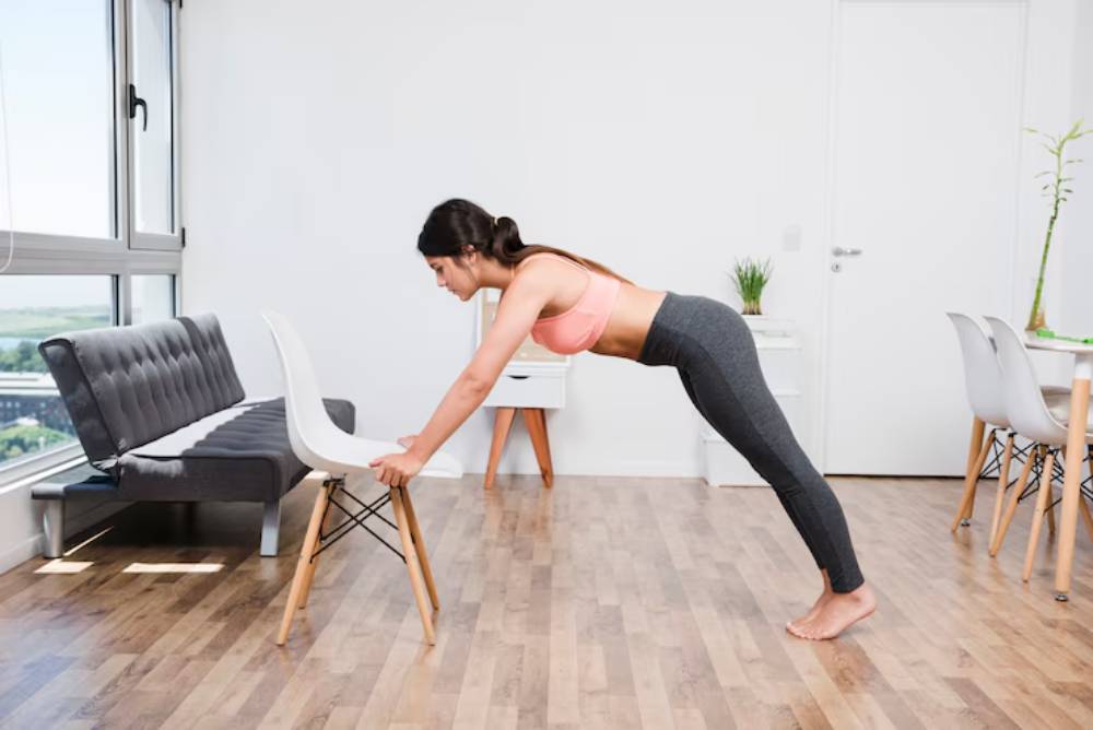 A woman in a pink sports bra and gray leggings performs an incline push-up against a chair in a bright, modern living space.
