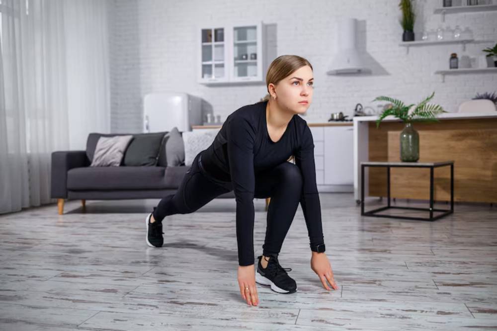Person in black athletic wear performing a lunge stretch in a modern living room with a sofa and minimalist decor.