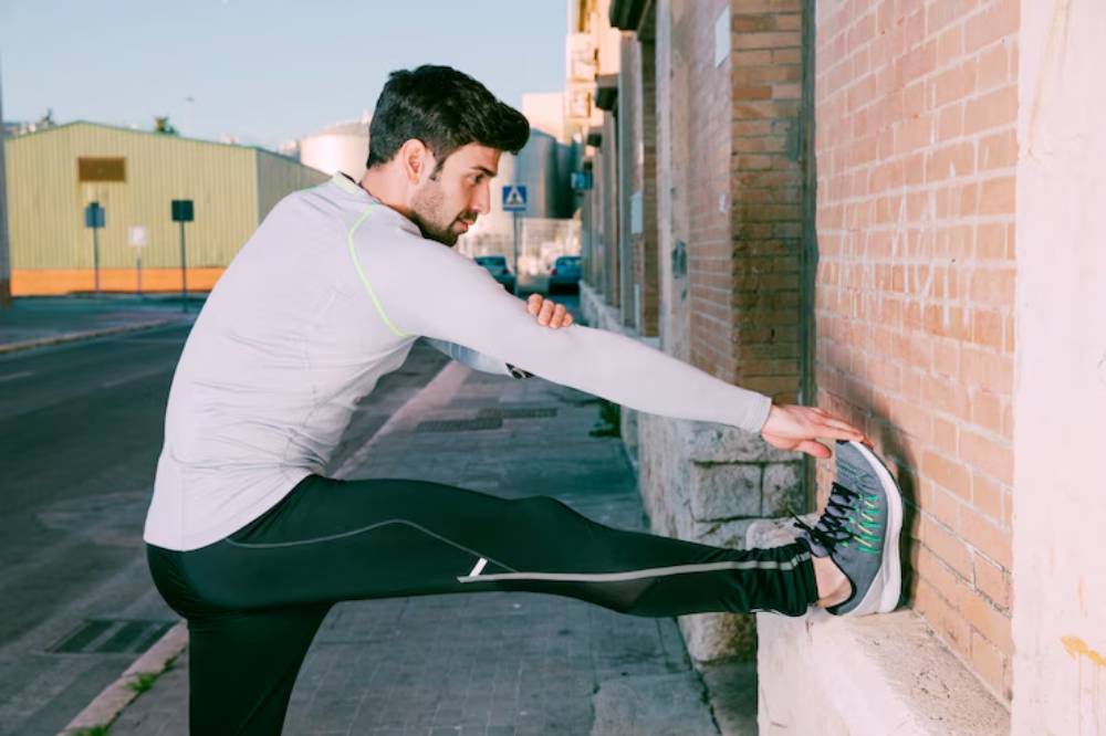 A person in athletic wear stretches their leg against a brick wall in an outdoor setting, preparing for a workout.