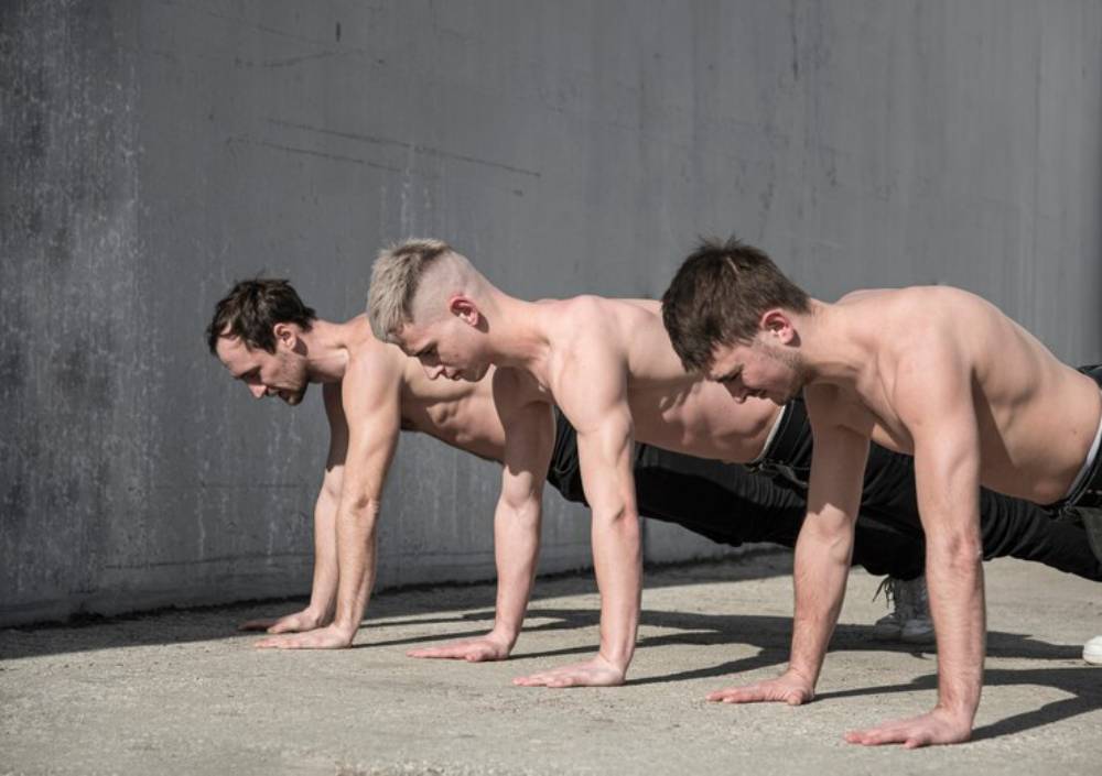 Three shirtless men perform push-ups in a line on a concrete surface, with a textured gray wall in the background.