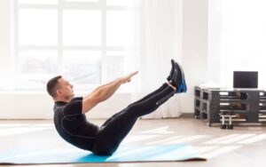 A person performs a V-sit exercise on a yoga mat in a bright room with large windows and workout equipment nearby.