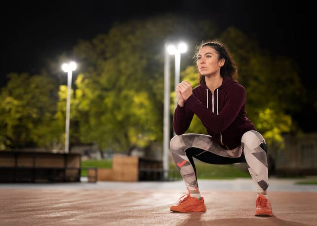 A person in athletic wear performs a squat on a nighttime outdoor court, surrounded by soft lighting and trees in the background.