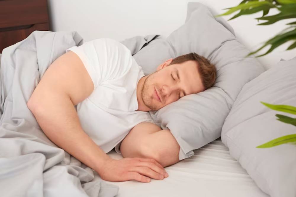 A person in a white t-shirt is peacefully sleeping on a bed, surrounded by soft gray bedding and pillows. A green plant is nearby.