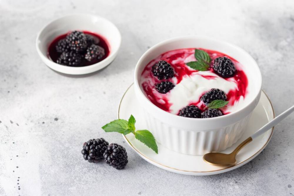 A creamy yogurt dessert topped with blackberries and raspberry sauce, garnished with mint, served in a white bowl with a gold spoon.