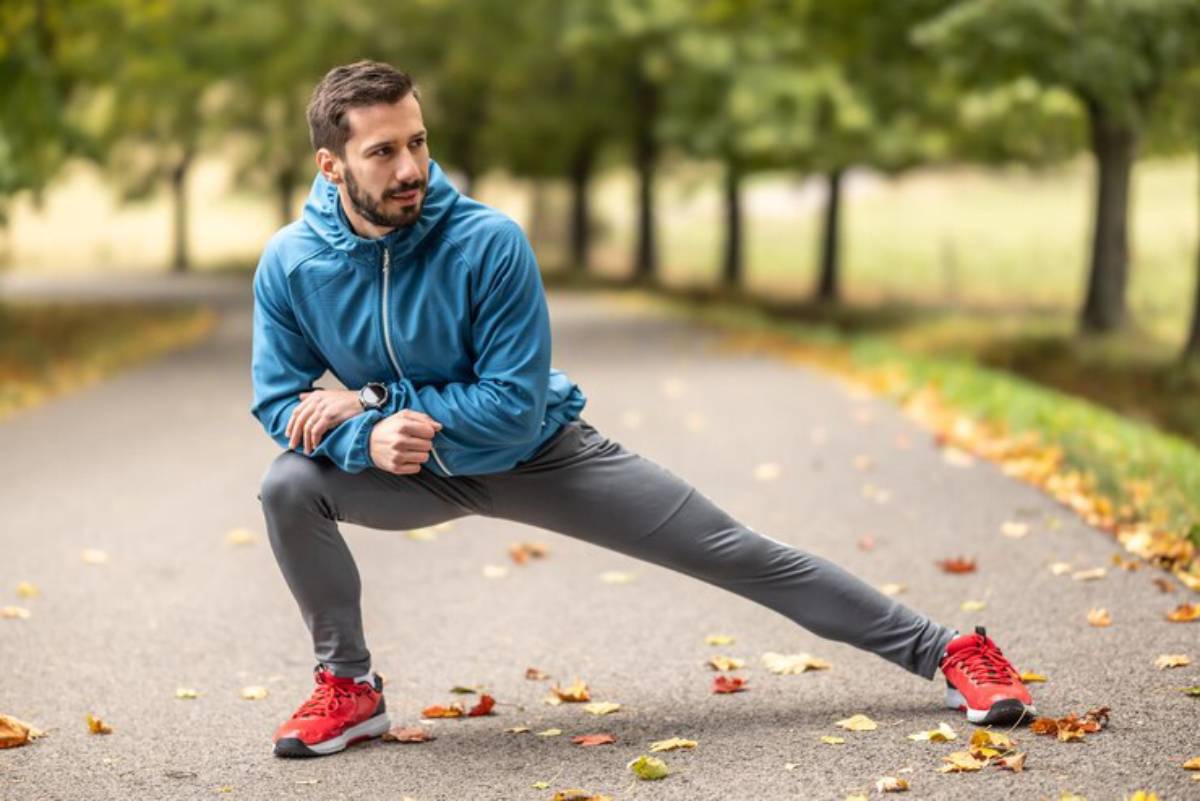 A person in a blue hoodie and gray leggings stretches in a park, surrounded by autumn leaves on the pathway.
