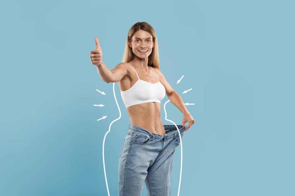 A person in a white sports bra and loose jeans holds their thumbs up, showcasing a significant weight loss achievement against a blue background.