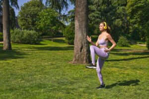A woman in purple athletic wear exercises in a sunny park, surrounded by lush green grass and trees.