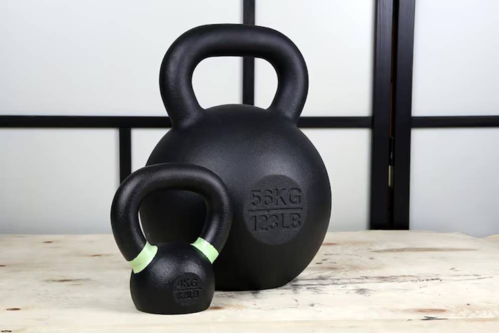 A large black kettlebell (56kg) stands next to a smaller kettlebell (8kg), both on a wooden surface, against a light background.