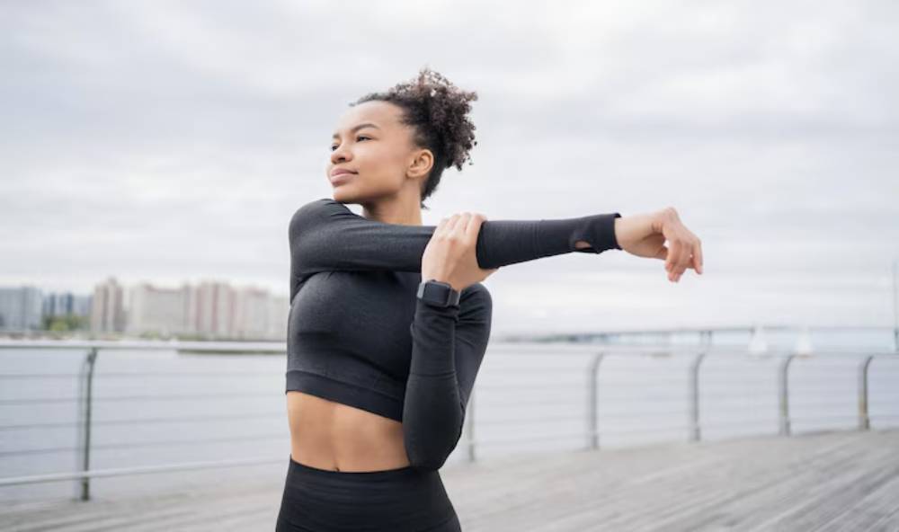 A woman in athletic wear stretches her arm on a waterfront deck, with a cloudy skyline and calm water in the background.