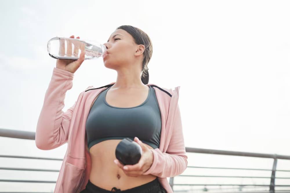 A person in sportswear drinks water from a bottle while holding a dumbbell, set against a bright, open sky.