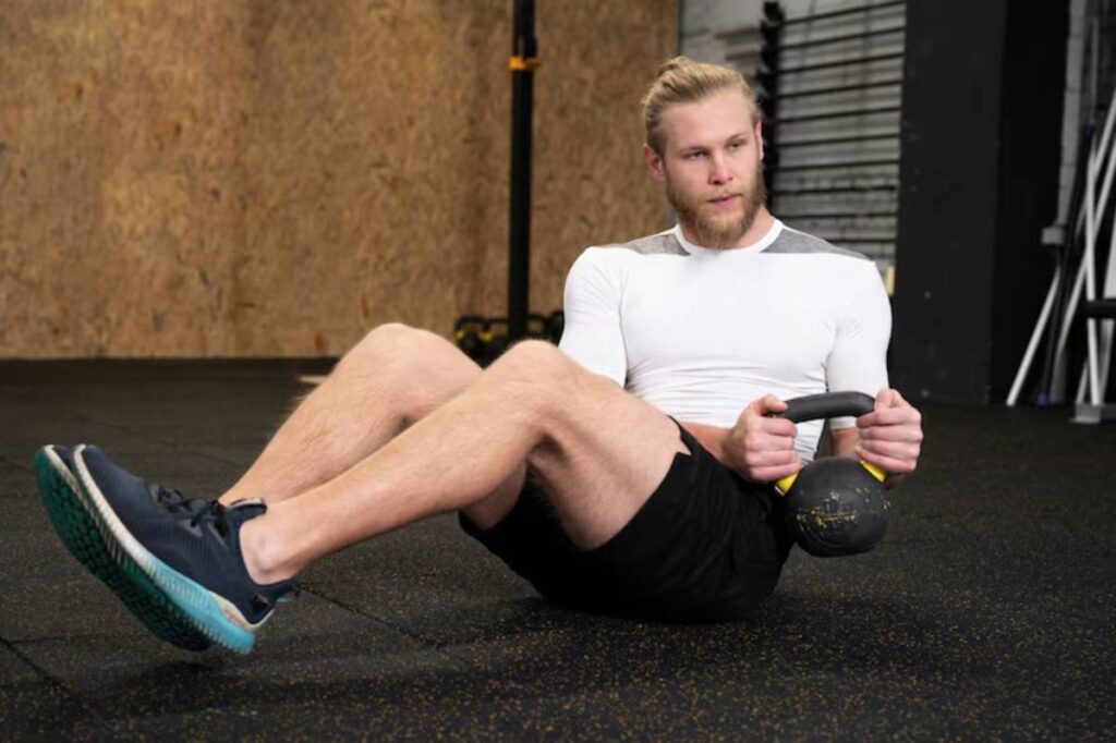 A person performs a seated kettlebell exercise on a gym floor, focusing on core strength and stability.