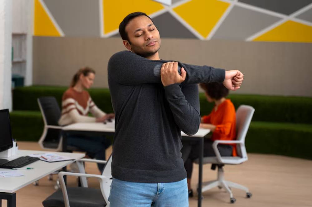 A person stretches their arm in a modern workspace, with colleagues engaged in conversation at nearby desks.