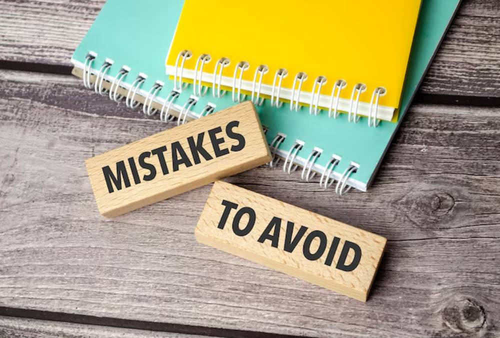 Wooden blocks reading 'MISTAKES' and 'TO AVOID' on a wooden surface, with colorful notebooks in the background.