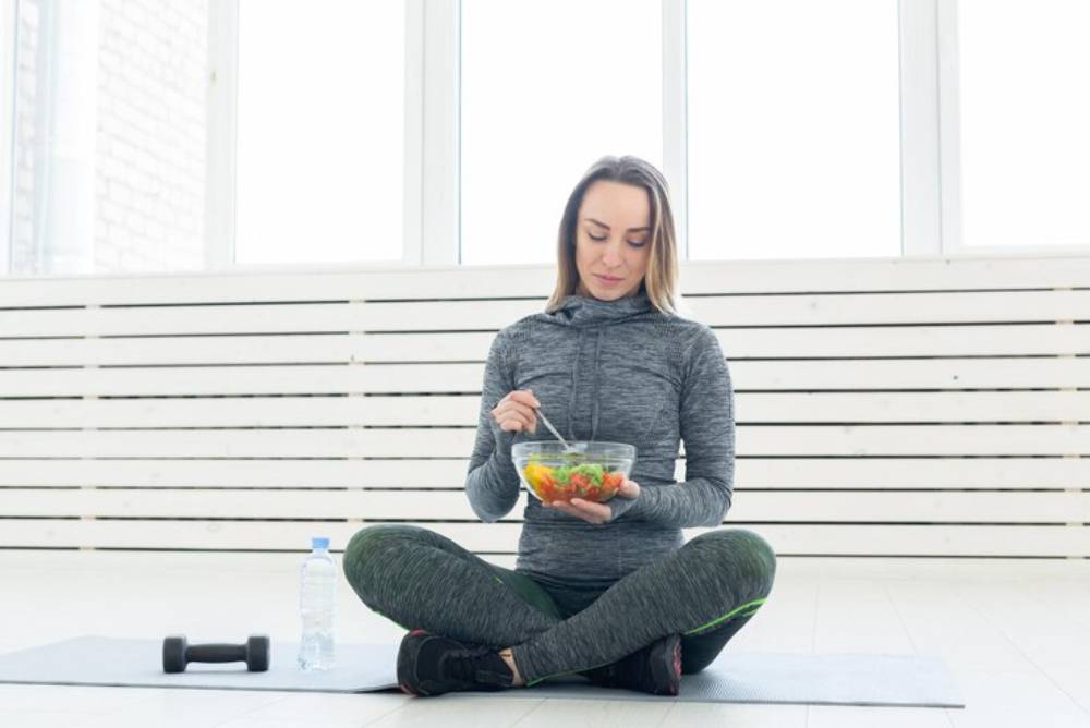 A person sits cross-legged on a yoga mat, enjoying a colorful salad in a bright, airy indoor space with a water bottle nearby.