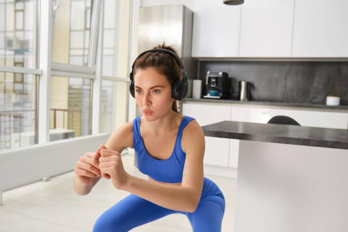 A person in a blue workout outfit is performing squats in a bright, modern kitchen while wearing headphones.