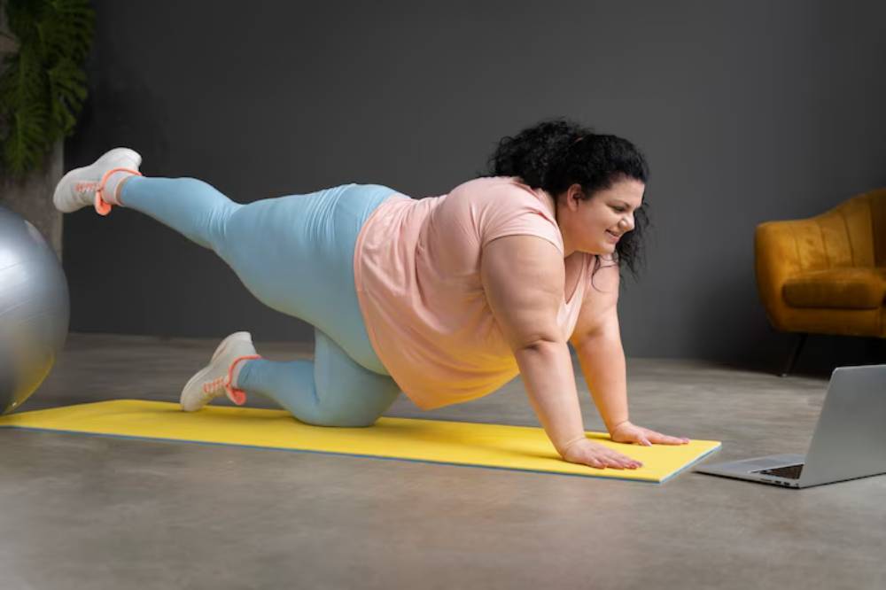 A fitness enthusiast performs an exercise on a yellow mat at home, with a laptop in front and a stability ball nearby.