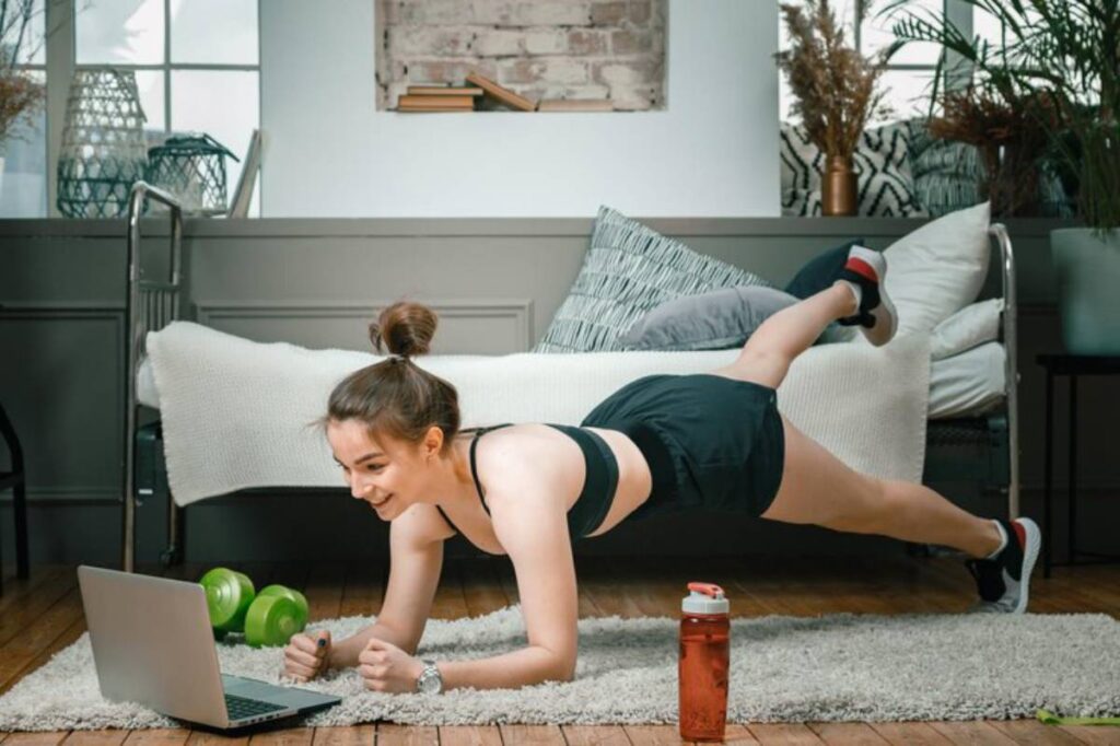 A person performs a plank exercise in a bright, stylish room, with weights and a laptop nearby, showcasing a home workout scene.