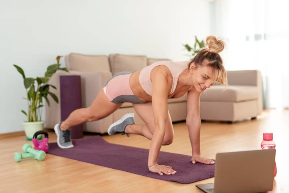 A woman exercises on a yoga mat at home, using a laptop for guidance, with dumbbells nearby and greenery in the background.