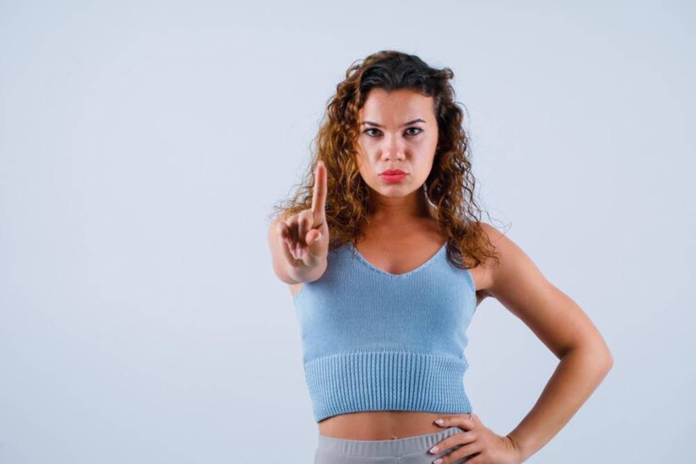 A woman with curly hair is posing against a plain background, holding up her index finger in a gesture of 'stop' or warning.