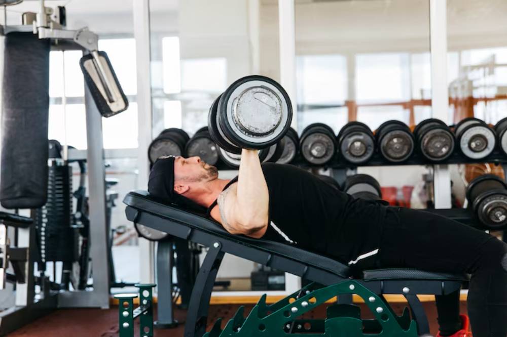 A man performs a dumbbell bench press in a gym, focused and lifting weights against a backdrop of exercise equipment.