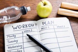 A workout journal with weekly plans, a pen, a green apple, and a water bottle on a wooden surface.