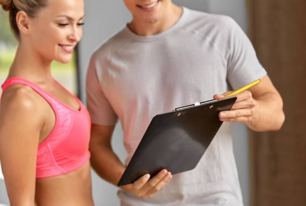 A fitness trainer smiles while reviewing workout plan on a clipboard, standing next to a client in athletic attire, inside a bright gym.