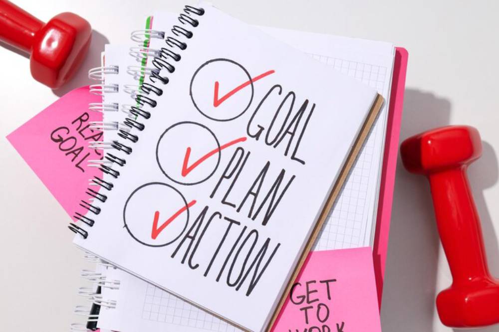 A notebook with 'GOAL,' 'PLAN,' and 'ACTION' checked off, surrounded by colorful sticky notes and a red dumbbell, symbolizing fitness planning.