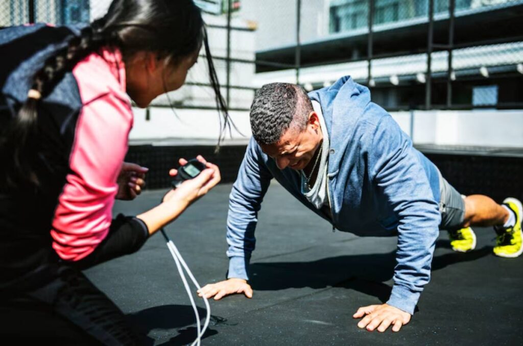 A person performs push-ups on a gym mat while another holds a timer and provides encouragement in a bright, outdoor workout space.