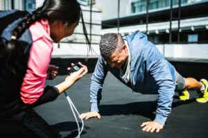 A person performs push-ups on a gym mat while another holds a timer and provides encouragement in a bright, outdoor workout space.