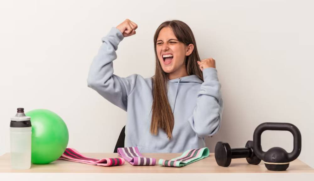 A woman in a gray hoodie celebrates fitness, surrounded by exercise equipment: dumbbells, a kettlebell, resistance bands, and a green ball.