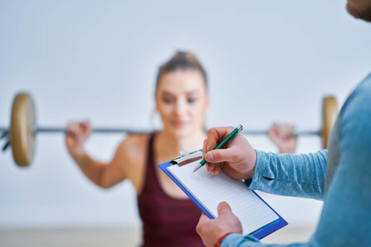 A person lifts a barbell while another takes notes on a clipboard, showcasing a fitness training session.