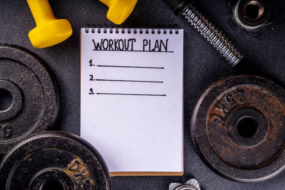 A notepad titled 'Workout Plan' with three blank lines, surrounded by weights and fitness equipment on a dark surface.