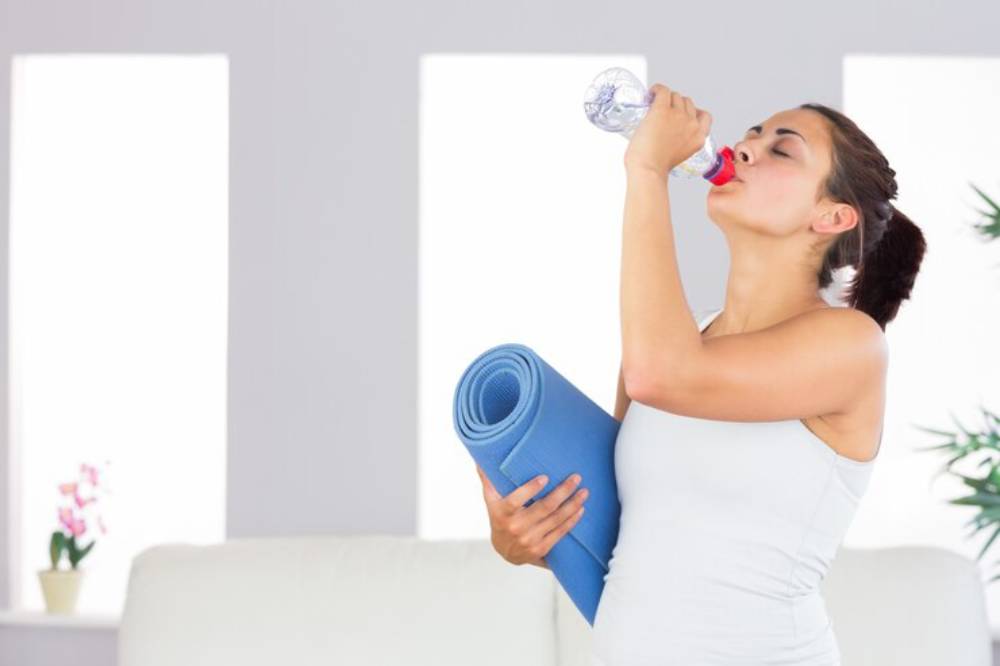 A woman in a white tank top holds a blue yoga mat and drinks water, surrounded by light-colored walls and decorative plants.