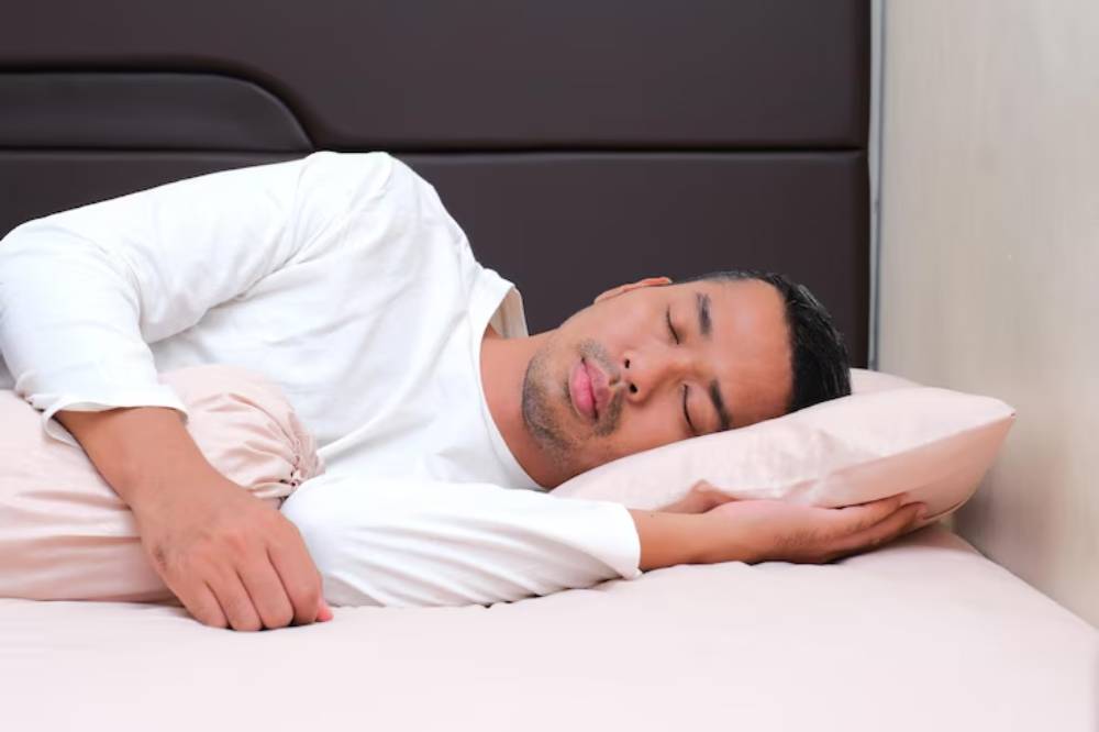 A person lies on a bed, sleeping on a pink pillow with their arm draped over it, against a neutral background.