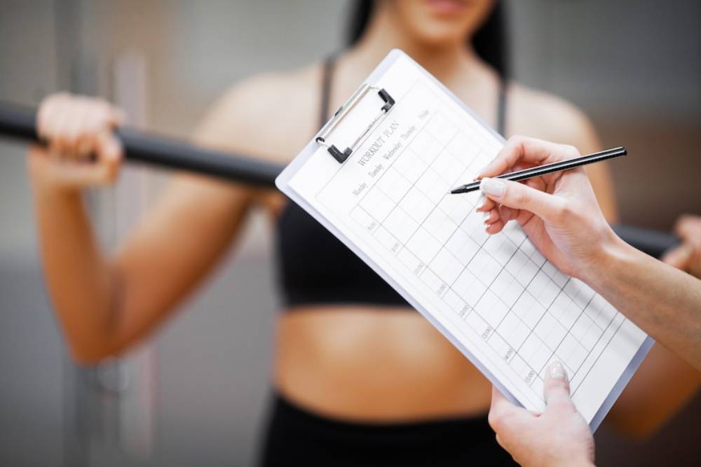 A person holds a clipboard with a workout plan while another prepares to lift a barbell, focusing on exercise and fitness.