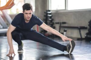 A young man in a gym performing a deep stretch, balancing on one leg while reaching towards his foot, doing warm-up before workout.