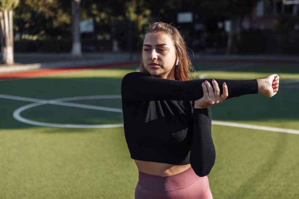 A woman in a black long-sleeve top stretches her arm on an outdoor sports field, surrounded by greenery and a blurred background.
