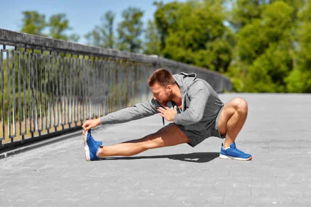 A person stretches on a paved surface with greenery in the background, wearing a gray hoodie, shorts, and blue athletic shoes.