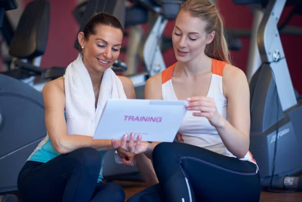 Two women in activewear sit on a gym floor, reviewing a training plan on a clipboard. Exercise equipment is visible in the background.
