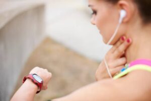 A person wearing a red smartwatch checks their fitness tracker while listening to music through white earbuds.