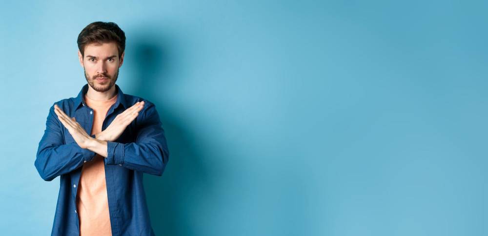 A person standing against a light blue backdrop, crossing their arms in a 'no' gesture, wearing a casual outfit.