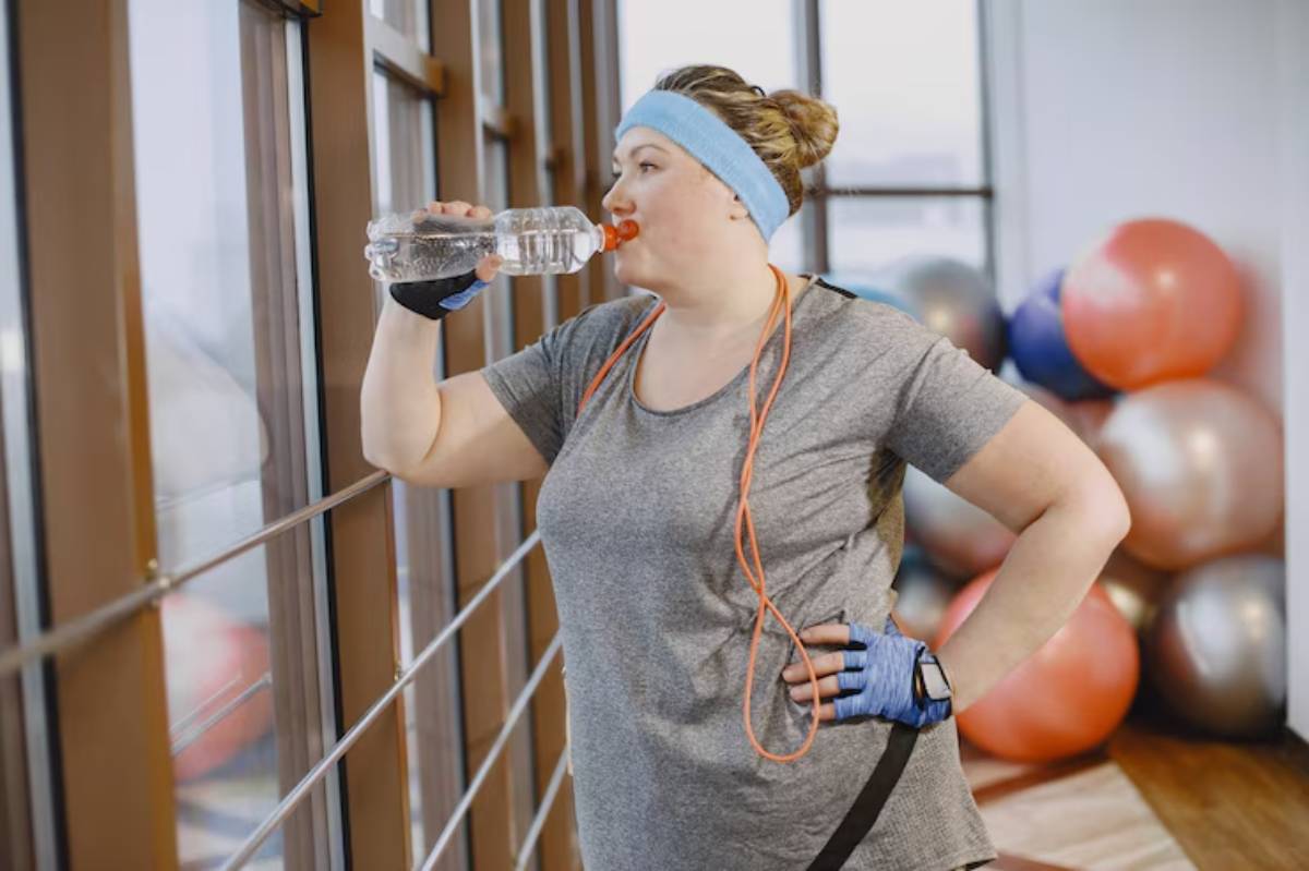 A woman with a ponytail, wearing athletic gear, takes a drink from a water bottle in a gym with exercise balls in the background.