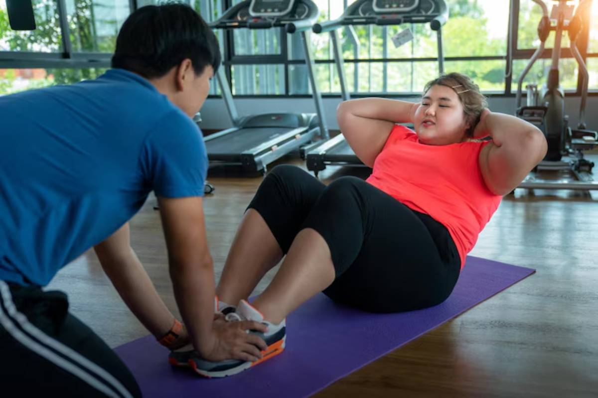 A person in a pink shirt performs crunches on a purple mat, while a trainer supports them in a bright gym setting with exercise equipment. 