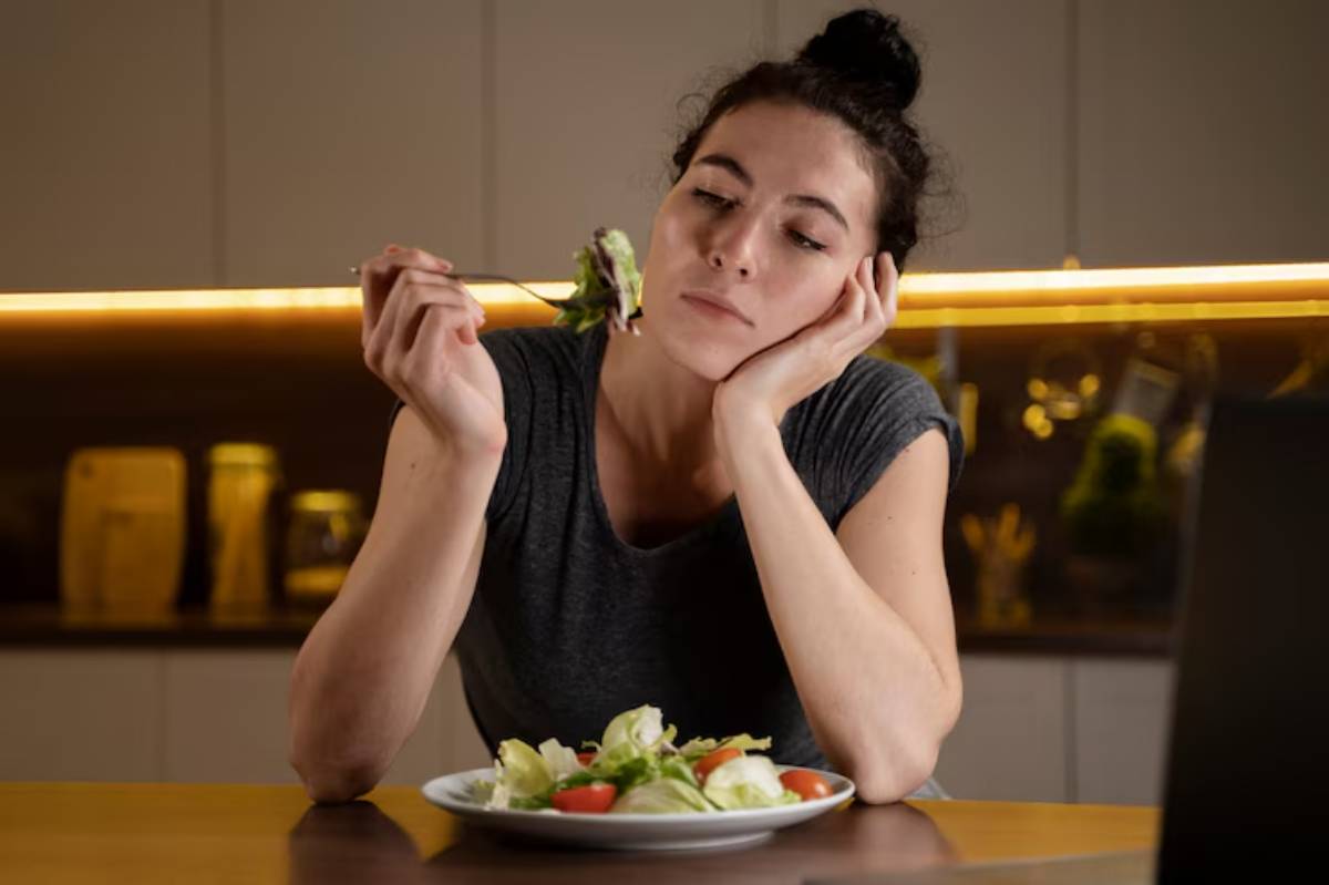 A person sits at a kitchen table, holding a fork with a salad, looking thoughtfully at the plate in front of them.