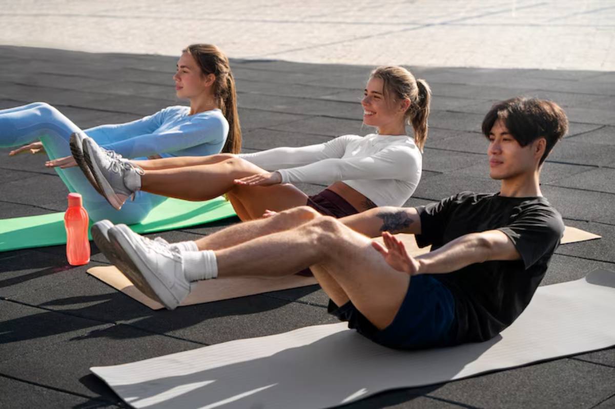 Three individuals practice ab workouts on yoga mats outdoors, with a water bottle placed nearby on the ground.