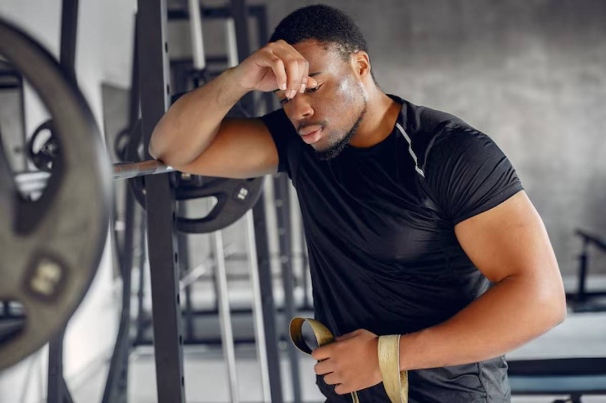 A muscular man in a black workout shirt leans against gym equipment, holding lifting straps, looking fatigued after overtraining.