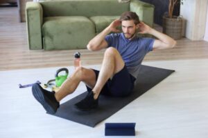 A person performs crunches on a yoga mat at home, surrounded by fitness equipment including a kettlebell and water bottle.