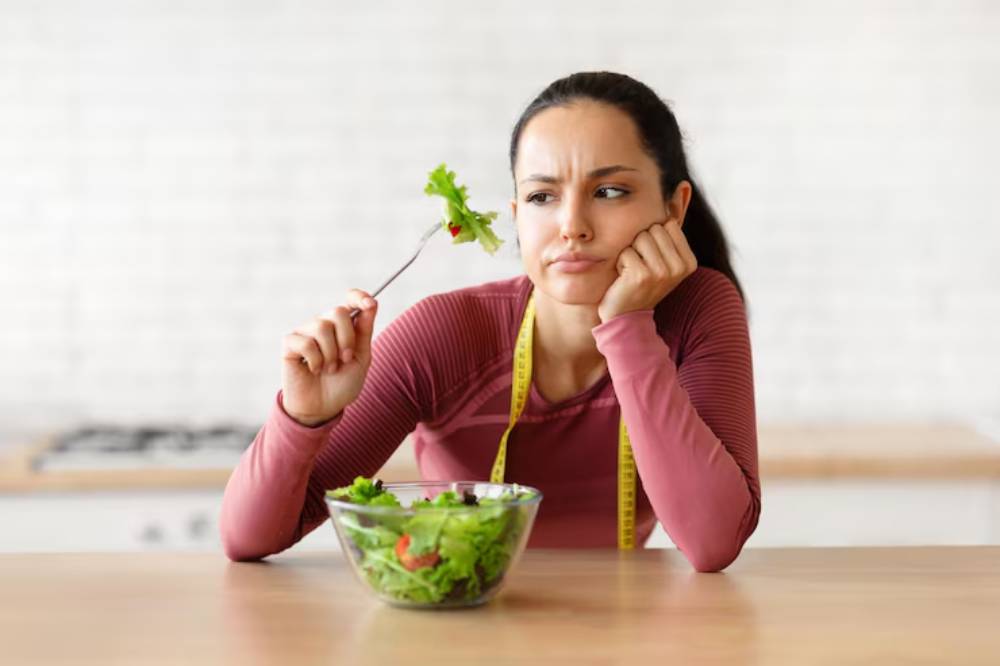 A person in a pink long-sleeve shirt holds a fork to a bowl of fresh salad, contemplating their meal in a bright kitchen.