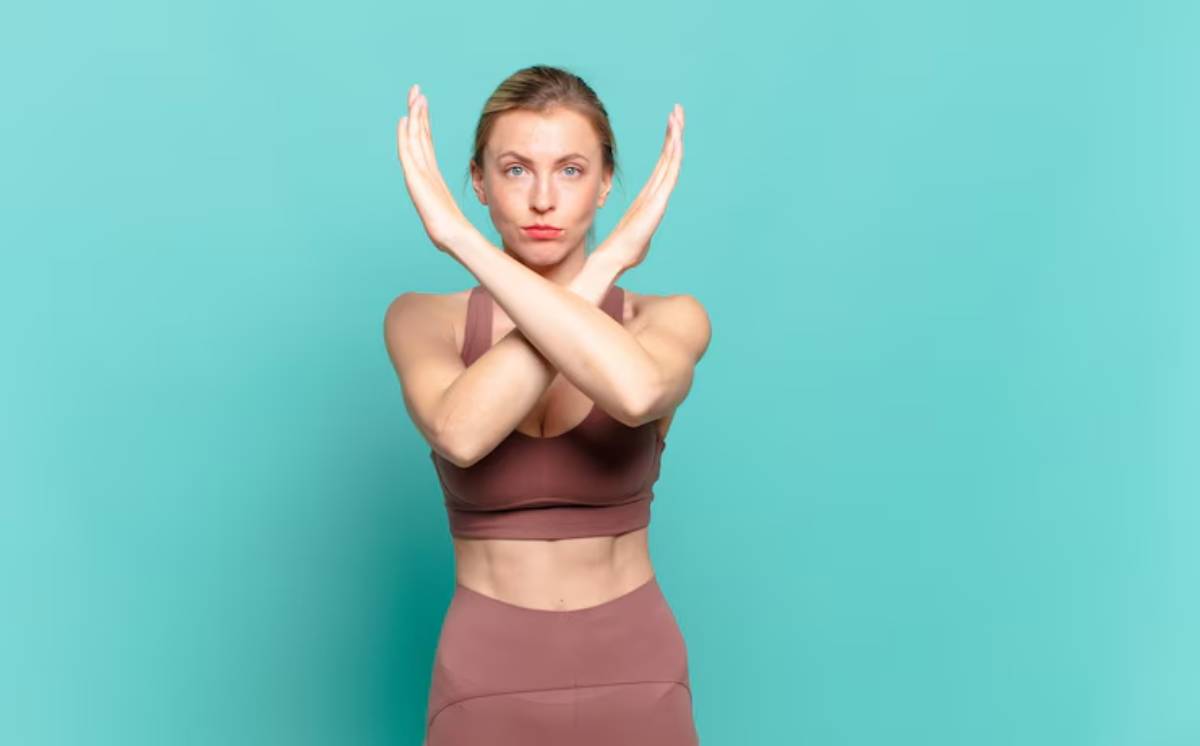 A woman in a brown sports bra and leggings poses against a turquoise background, crossing her arms in front of her.