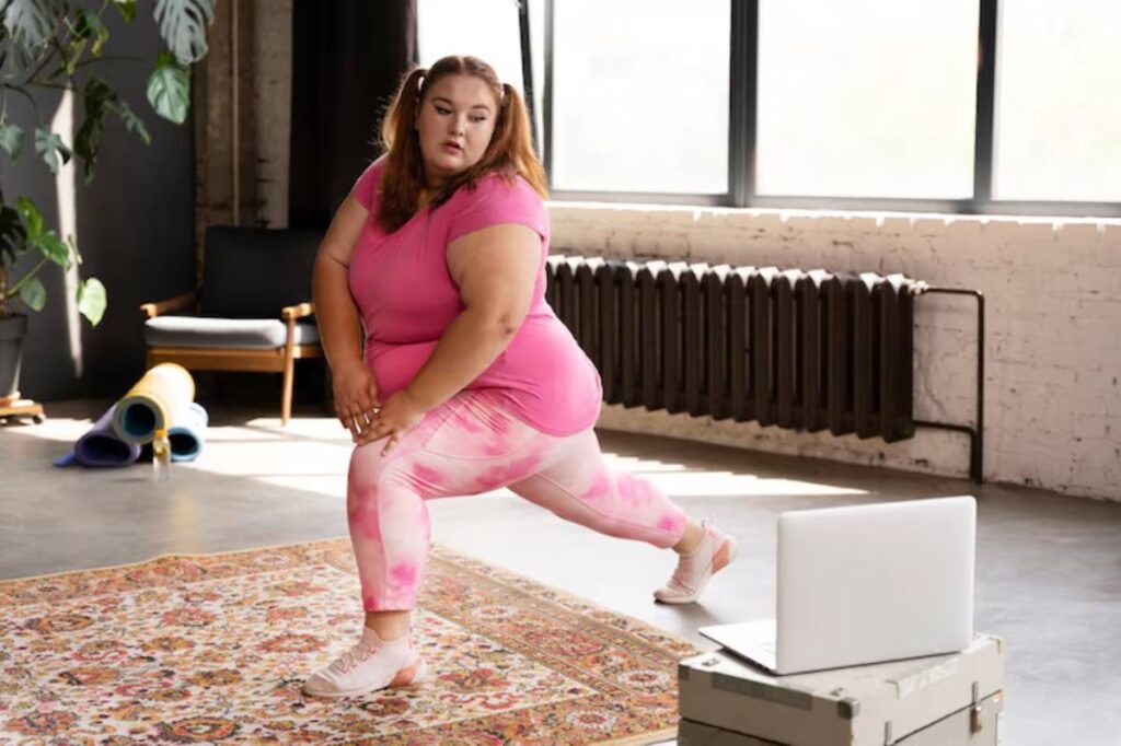 A person in a pink workout outfit performs a lunge at home, with a laptop on a crate and yoga mats nearby in a spacious room.